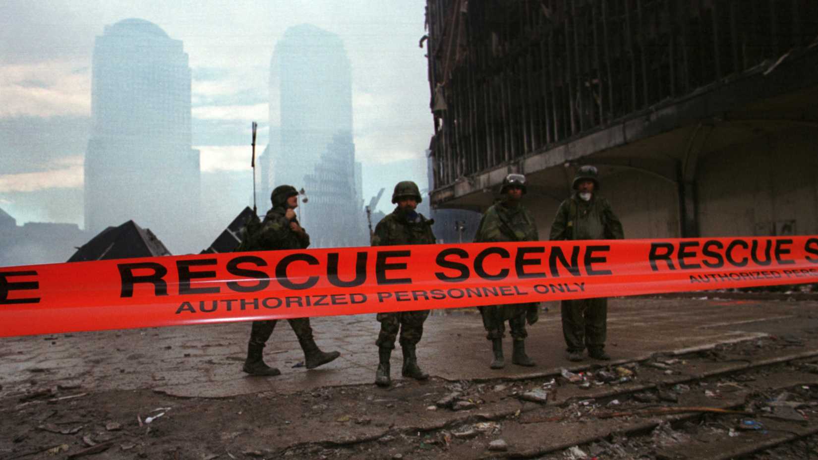Four soldiers stand behind orange “Rescue Scene” tape at Ground Zero, with debris underfoot and buildings obscured by haze.