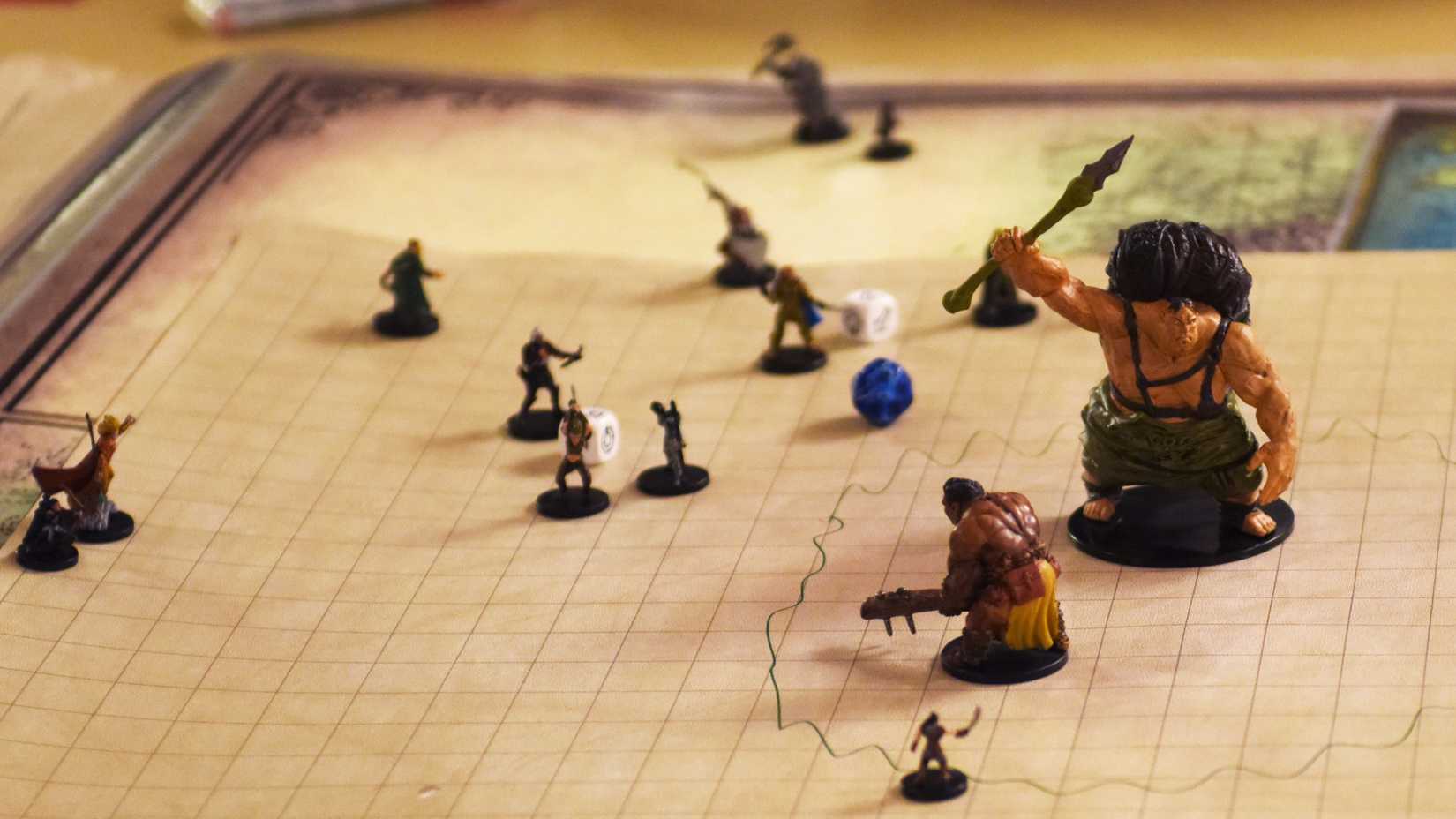Fantasy miniatures and dice stand on a gridded battle map, with a large spear-wielding figure in the foreground.
